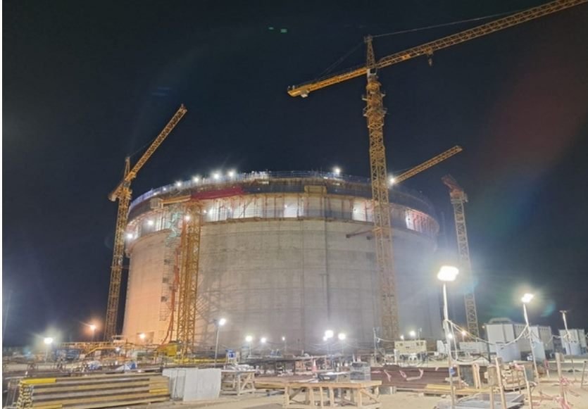 Illuminated LNG storage tank under construction at night, flanked by two tower cranes